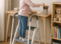 Young girl standing on a three-step white and gray toddler stool, reaching for colored pencils on a wooden desk in a cozy study corner with acoustic panels and a bookshelf featuring a teddy bear and children's books.